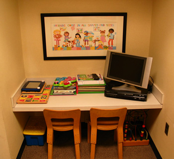 The image shows an interior space with a small desk area featuring a computer monitor, keyboard, mouse, and books, along with two chairs. Above the desk hangs a framed poster with a colorful illustration of children engaged in various activities.