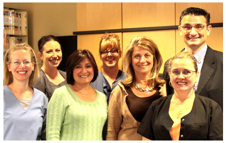 The image shows a group of seven individuals posing together for a photograph. They are standing in front of a backdrop, likely an event setting, with a person at the far left wearing glasses and a bright green top, followed by a woman in a blue shirt, then another individual partially visible on the right side of the frame. The group appears to be a mix of genders and ages, all smiling and looking directly at the camera.