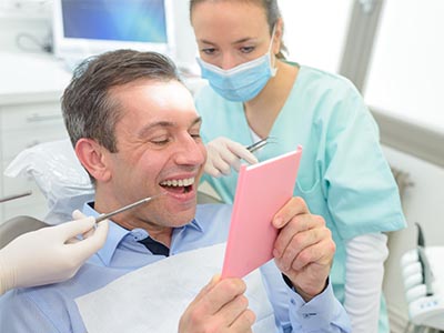 A man with a toothy grin is looking at a pink card he holds up while sitting in a dental chair, surrounded by dental equipment and a smiling female dental professional.