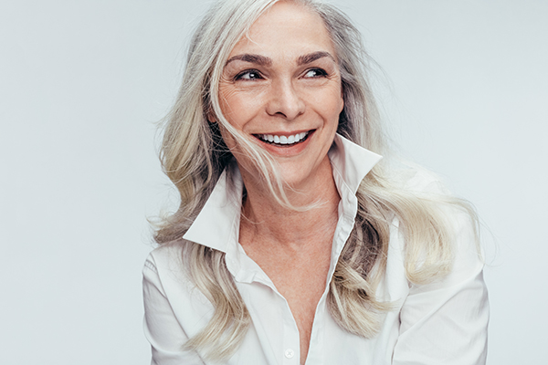 The image shows a woman with short blonde hair smiling at the camera, wearing a white top and sitting against a light background.