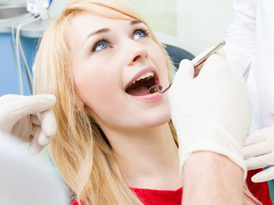 A woman sitting in a dental chair with a dental professional performing an oral examination, wearing gloves and a surgical mask, while the woman has her mouth open.