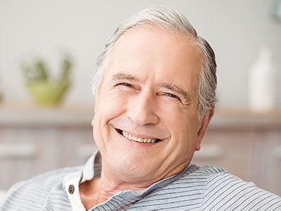 The image shows a smiling older man with gray hair, wearing a blue shirt, sitting comfortably in a chair indoors, with a warm, inviting ambiance.