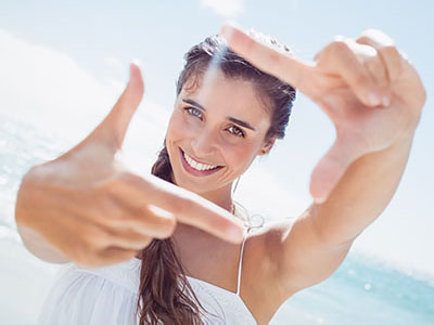 The image features a young woman with long hair smiling at the camera, holding up her index finger as if framing a photo, against a bright blue sky background.